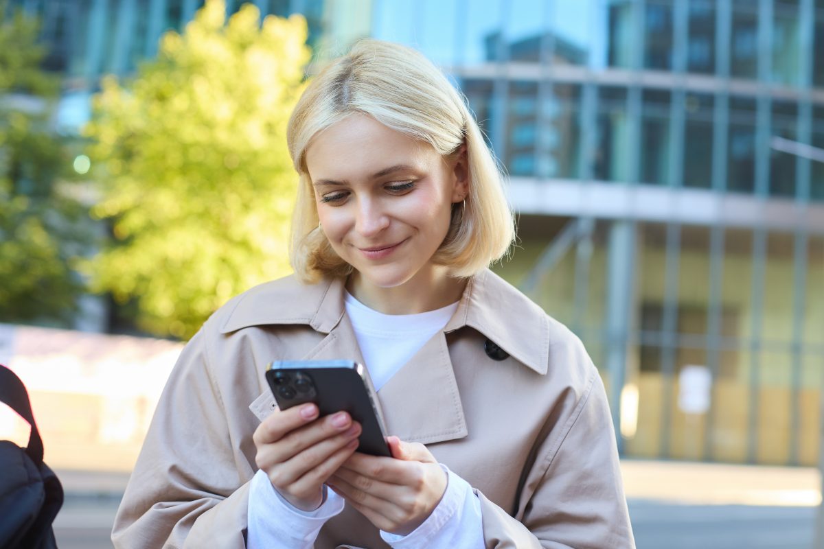 Close up portrait of smiling female model on street, sitting on bench with mobile phone, looking at smartphone screen, messaging with friend while waiting outside